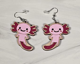 May include: A pair of pink and red axolotl earrings. The axolotls are smiling and have small, black eyes. The earrings are made of wood and have silver hooks.