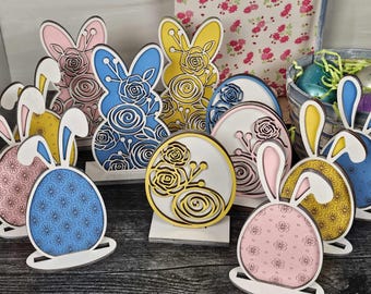 May include: A set of wooden Easter decorations featuring bunnies and eggs. The decorations are painted in various colors, including pink, blue, yellow, and white. Each decoration has a floral design and is standing on a wooden base.