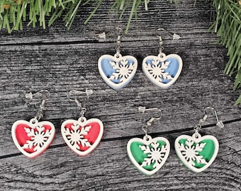May include: Heart-shaped earrings in red, blue, and green with a white snowflake design. The earrings have a white border and silver-colored hooks. The earrings are displayed on a dark wooden surface with evergreen branches.