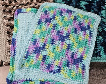 May include: Close-up of several handmade crochet potholders. The potholders feature various color schemes, including teal, purple, blue, and green. The crochet work is detailed, with visible stitch patterns and a textured appearance. The potholders are square-shaped and appear to be made of cotton yarn.