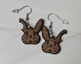 May include: A pair of wooden earrings shaped like bunnies with surprised expressions. The earrings are brown and have a wood grain texture. The bunny ears are slightly pointed and the bunnies have small, round eyes.