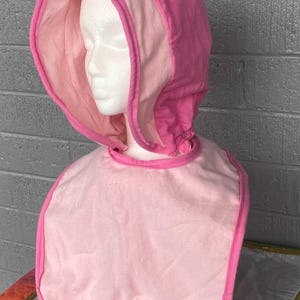 May include: A pink and light pink bonnet with a bib-like front, displayed on a mannequin head. The bonnet has a hood with a darker pink trim and lining. The bib is a lighter pink with a darker pink trim.