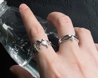 May include: Two silver rings on a finger. One ring is a simple band with a small pearl accent. The other ring is a more intricate design with a wave-like pattern.