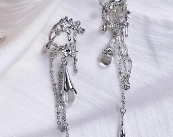 May include: A pair of silver ear cuffs with dangling chains and teardrop-shaped charms. The cuffs are designed with a delicate, intricate pattern that resembles dripping water.