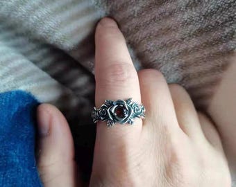 May include: A silver-toned ring featuring a rose design with a central red gemstone. The ring has detailed floral elements and is worn on a finger. The ring's design is intricate and detailed, with a vintage or gothic aesthetic.