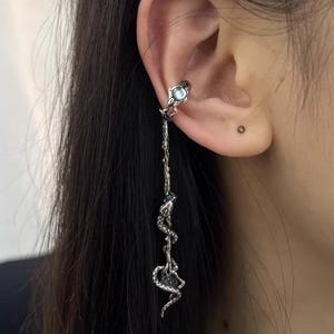 May include: Silver ear cuff with a dangling snake charm and a blue gemstone. The snake is coiled and has a detailed design.