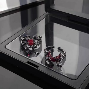 Peut inclure: Deux bagues en argent avec des accents de pierres précieuses rouges. Les bagues présentent des motifs complexes, l'une avec une forme de croissant de lune et l'autre avec un motif de vigne tourbillonnant.