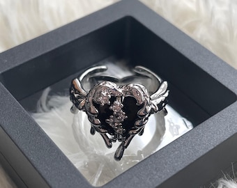 Könnte beinhalten: Ein silberner Ring mit einem schwarzen Herzdesign und klaren Strasssteinen. Das Herz tropft mit Silber und hat an beiden Seiten Flügel.