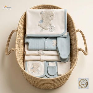Peut inclure: Un panier en osier tressé contenant un ensemble de vêtements et accessoires pour bébé dans des tons crème et bleu clair. Les articles incluent une couverture, un gilet à manches longues, un body et des moufles, ornés de motifs brodés.