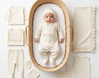 May include: A newborn baby in a cream-colored outfit lies in a woven basket, surrounded by matching clothing items. The baby wears a onesie with a bow tie and a hat. Additional items include a blanket, bib, pants, shirt, and a pair of mittens.