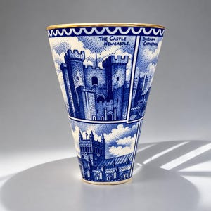 May include: A conical ceramic vase featuring a blue and white transferware pattern of The Castle Newcastle and Durham Cathedral. The rim is accented with a gold band and a scalloped blue border. The design includes stippled clouds and architectural details.