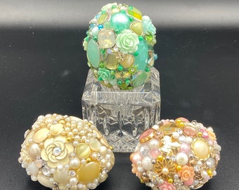 May include: Three decorative eggs covered in various beads, rhinestones, and small flowers. The eggs are in shades of green, white, and pink.