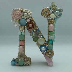May include: A decorative letter N made of a variety of materials, including beads, buttons, and flowers. The letter is in a light blue, pink, and green color scheme.