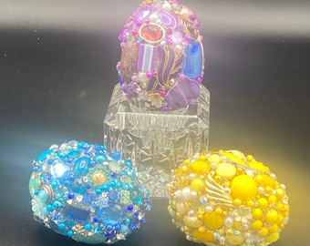 May include: Three decorative eggs covered in colorful beads, rhinestones, and other embellishments. The eggs are purple, blue, and yellow.