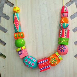 May include: A colorful, handcrafted necklace with a variety of painted wooden beads. The beads are in various shapes, including round, cylindrical, and geometric, and feature bright colors like pink, orange, green, and turquoise. The necklace is displayed on a wooden stand.