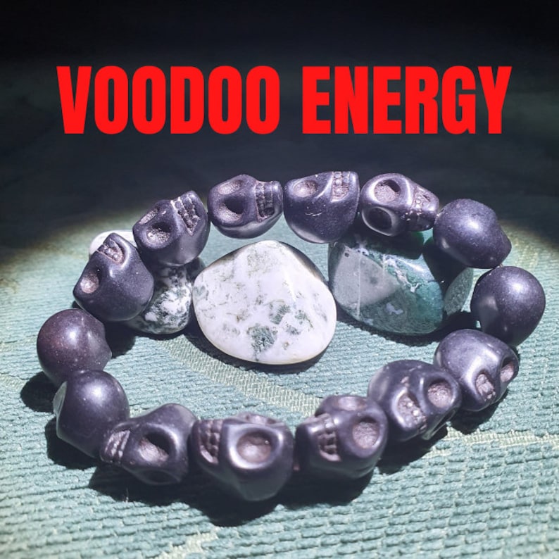 May include: A black skull bead bracelet with the words "VOODOO ENERGY" in red text. The bracelet is on a green fabric surface with two green and white stones.