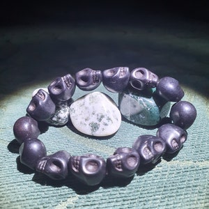 May include: A black skull bead bracelet with a green and white agate stone in the center. The bracelet is made of black beads that are shaped like skulls.