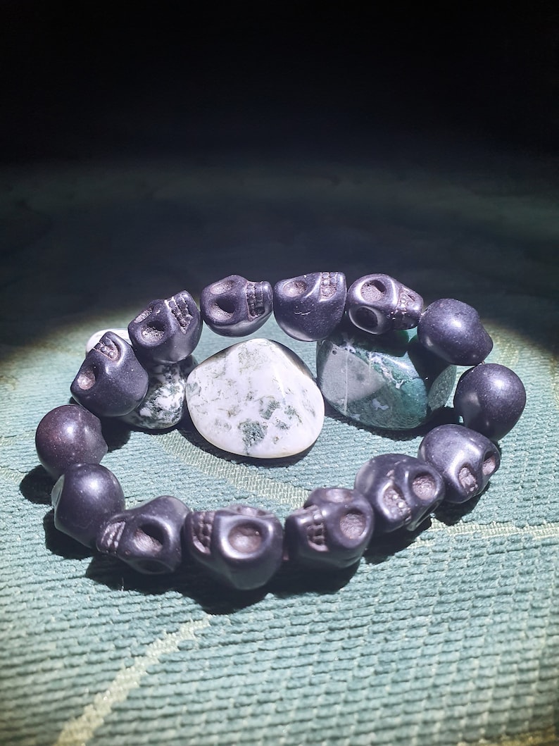 May include: A black skull bead bracelet with a green and white agate stone in the center. The bracelet is made of black beads that are shaped like skulls.