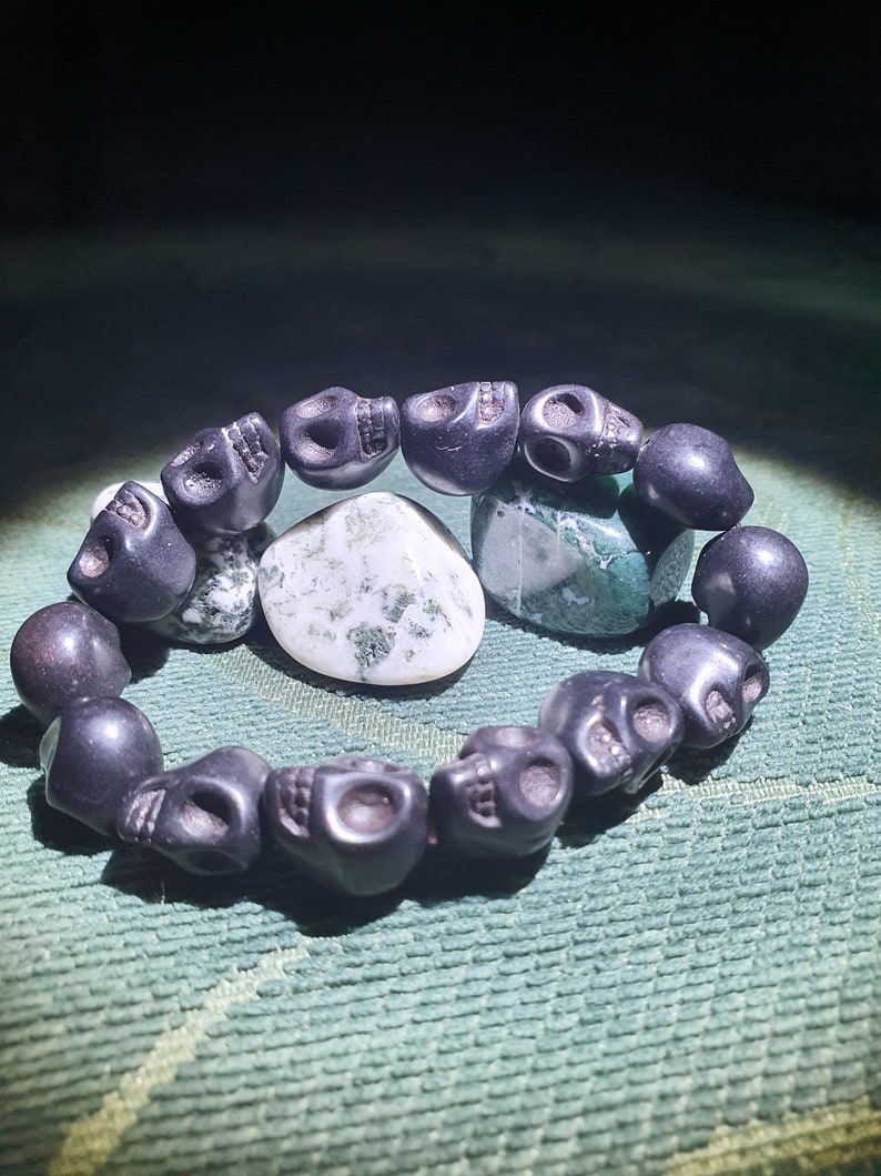 May include: A black skull bead bracelet with a green and white agate stone. The bracelet is made of black beads that are shaped like skulls. The agate stone is in the center of the bracelet.