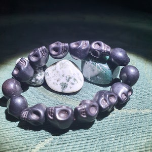May include: A black skull bead bracelet with a green and white agate stone. The bracelet is made of black beads that are shaped like skulls. The agate stone is in the center of the bracelet.
