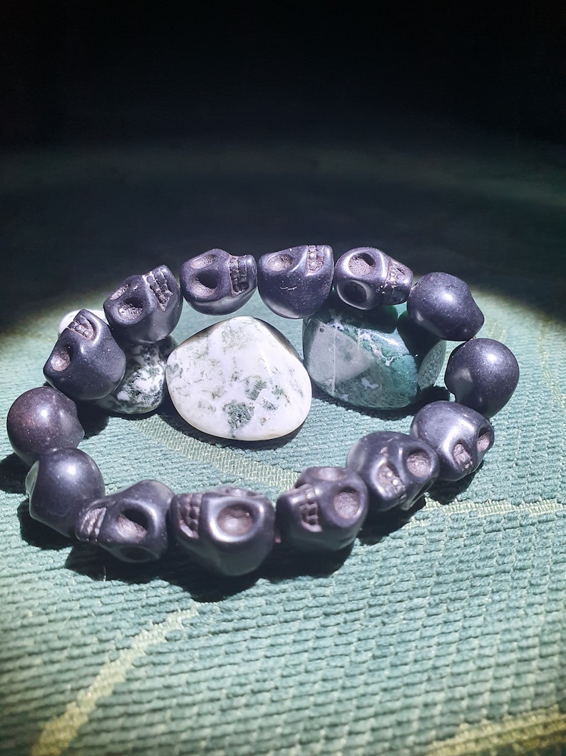 May include: A black skull bead bracelet with a green and white agate stone. The bracelet is made of black beads that are shaped like skulls. The agate stone is in the center of the bracelet.