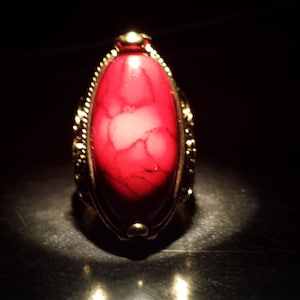 Könnte beinhalten: Ein goldfarbener Ring mit einem ovalen, roten Edelstein. Der Edelstein hat ein marmoriertes Aussehen mit helleren roten und weißen Adern. Der Ring ist vor einem dunklen Hintergrund platziert, wobei sich das Licht auf der Oberfläche spiegelt.