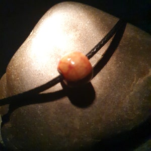 Peut inclure: Gros plan d'une perle rouge et blanche sur un collier en cordon noir. La perle est ronde et a une surface lisse. Le collier est posé sur une roche grise.