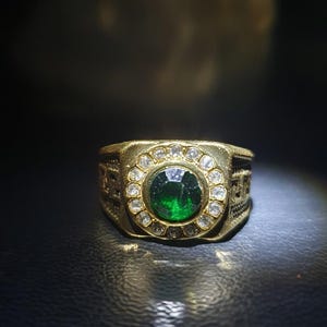 May include: A gold-toned ring featuring a large, round, emerald-green gemstone at the center, surrounded by a halo of smaller, clear stones. The ring's band has intricate detailing. The ring is displayed on a dark surface.