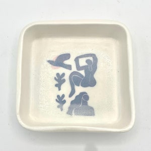 May include: A square, off-white ceramic dish with rounded corners. The interior features a design of abstract figures and shapes in blue and pink. The dish is ideal for holding small items.