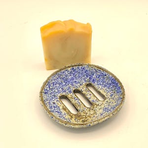 Könnte beinhalten: Handgefertigte Keramik-Seifenschale mit gesprenkelter blau-beiger Glasur und drei Abflussöffnungen. Ein rechteckiger Block hellorangener Seife liegt hinter der Schale. Ein funktionales Badezimmer-Accessoire.