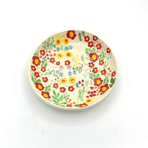 May include: A circular, cream-coloured ceramic dish featuring a hand-painted floral pattern. The flowers are red, yellow, and blue, with green foliage. The dish has a slightly raised rim and is set against a white backdrop.
