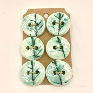 May include: Six ceramic buttons with a leaf design. The buttons are round, with a light green and white glaze and a dark green leaf pattern. Each button has two holes and is attached to a brown cardboard backing with twine.