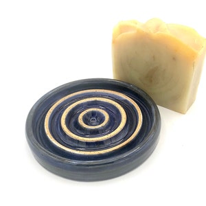 May include: A dark blue ceramic soap dish with a spiral design and a light-coloured bar of soap. The soap dish has a circular shape with concentric ridges. The soap is a light tan colour and has a textured surface.