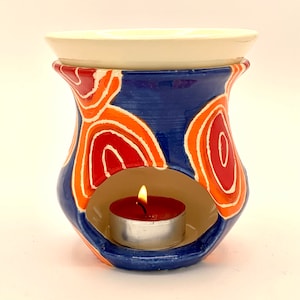 Puede incluir: Un difusor de aceites esenciales de cerámica con base azul, adornado con diseños abstractos en naranja y rojo. Cuenta con una parte superior blanca y una abertura circular para una vela de té encendida. La llama de la vela es visible.