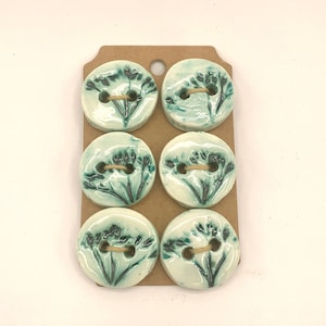 May include: Six ceramic buttons with a floral design in shades of teal and green, attached to a brown tag. Each button has two holes and a twine thread. The buttons are round and have a glossy finish.