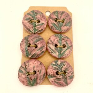 May include: Six round, ceramic buttons with a pink and green leaf design. Each button has two holes and is attached to a brown tag with twine. These buttons are a unique craft supply.