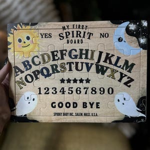 Pu&ograve; includere: Un puzzle di tavola Ouija in legno con il testo "MY FIRST SPIRIT BOARD". La tavola presenta l'alfabeto, i numeri e le parole "YES", "NO" e "GOOD BYE". Include illustrazioni di un sole sorridente, una luna crescente e due fantasmi.