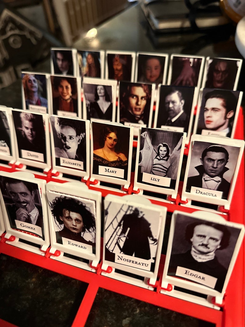 Peut inclure: Un plateau de jeu avec des cartes blanches repr&eacute;sentant des personnages de films d'horreur. Les cartes sont dispos&eacute;es en rang&eacute;es et en colonnes, et chaque carte contient une image d'un personnage et son nom. Parmi les personnages, on trouve Gomez, Edward, Nosferatu, Edgar, Dracula, Lily, Mary, Elizabeth, David, et bien d'autres.