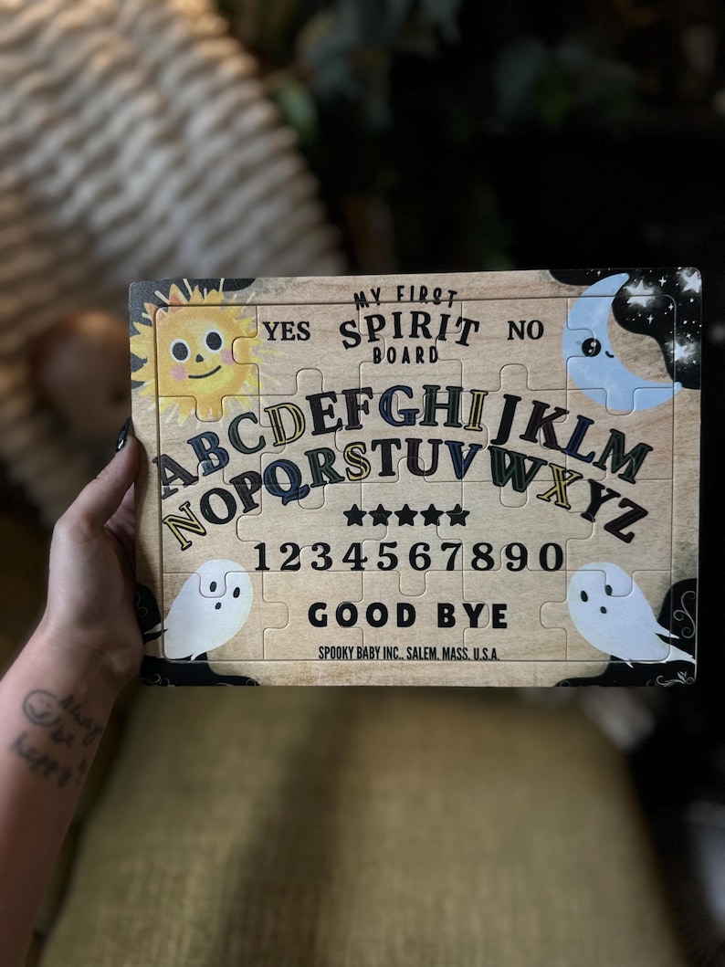 Pu&ograve; includere: Un puzzle di tavola Ouija in legno con il testo "MY FIRST SPIRIT BOARD". La tavola presenta l'alfabeto, i numeri e le parole "YES", "NO" e "GOOD BYE". Include illustrazioni di un sole sorridente, una luna crescente e due fantasmi.