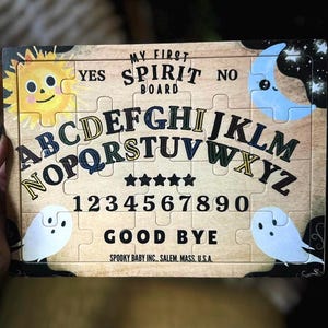 Pu&ograve; includere: Un tabellone di puzzle in legno con la scritta "MY FIRST SPIRIT BOARD". Il tabellone presenta lettere, numeri e le parole "YES", "NO" e "GOOD BYE". Ha illustrazioni di sole, luna e fantasmi.