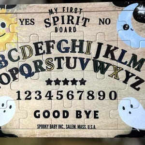 Pu&ograve; includere: Un puzzle in legno a tema spettrale. La tavola presenta il testo "MY FIRST SPIRIT BOARD", lettere, numeri e le parole "YES", "NO" e "GOOD BYE". I pezzi del puzzle includono illustrazioni di un sole sorridente, una luna e fantasmi.