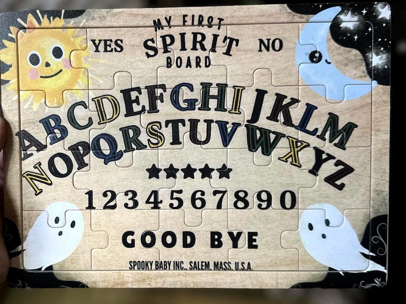 Pu&ograve; includere: Un puzzle in legno a tema spettrale. La tavola presenta il testo "MY FIRST SPIRIT BOARD", lettere, numeri e le parole "YES", "NO" e "GOOD BYE". I pezzi del puzzle includono illustrazioni di un sole sorridente, una luna e fantasmi.