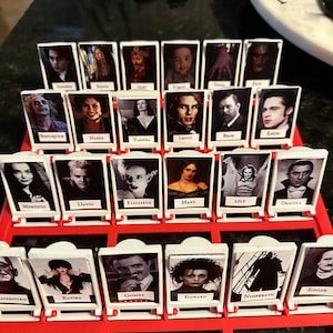 Peut inclure: Un plateau de jeu avec des tuiles en plastique blanc repr&eacute;sentant des personnages de films d'horreur. Les tuiles sont dispos&eacute;es en grille avec les noms des personnages imprim&eacute;s sous chaque image. Les noms incluent Beetlejuice, Nancy, Vampira, Lestat, Bram, Louis, Morticia, David, Elizabeth, Mary, Lily, Dracula, Frankenstein, Elvira, Gomez, Edward, Nosferatu et Edgar.