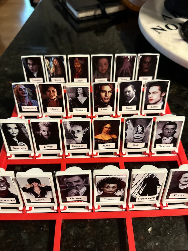 Peut inclure: Un plateau de jeu avec des tuiles en plastique blanc repr&eacute;sentant des personnages de films d'horreur. Les tuiles sont dispos&eacute;es en grille avec les noms des personnages imprim&eacute;s sous chaque image. Les noms incluent Beetlejuice, Nancy, Vampira, Lestat, Bram, Louis, Morticia, David, Elizabeth, Mary, Lily, Dracula, Frankenstein, Elvira, Gomez, Edward, Nosferatu et Edgar.