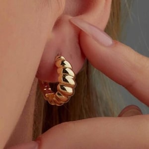 May include: Gold hoop earrings with a textured, ridged design. The earrings are worn in a person's ear.