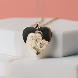 May include: A heart-shaped pendant necklace with a gold chain. The pendant features a black and white engraved image of a couple. The necklace is displayed on a white surface with a pink background.