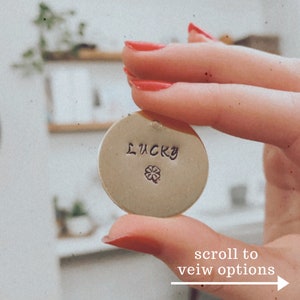 Pode incluir: Um amuleto de metal dourado com a palavra "LUCKY" gravada nele. Um trevo de quatro folhas também está gravado no amuleto.