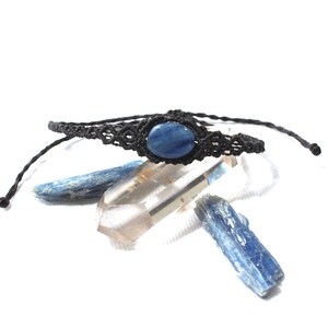 May include: A black macrame bracelet featuring a blue gemstone. The bracelet is shown on a white surface with blue and clear crystals.