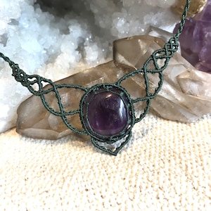 May include: A handcrafted macrame necklace featuring a large, polished amethyst stone. The necklace is made with dark green cord, showcasing intricate knotting patterns around the central stone. The amethyst is a deep purple color, set against a neutral background.