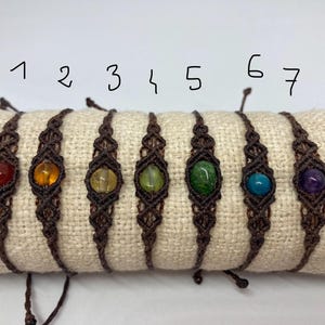 Peut inclure: Sept bracelets en macramé marron avec des perles de pierres précieuses de différentes couleurs. Les perles sont rouge, orange, jaune, verte, bleu sarcelle et violette. Les bracelets sont numérotés de 1 à 7.